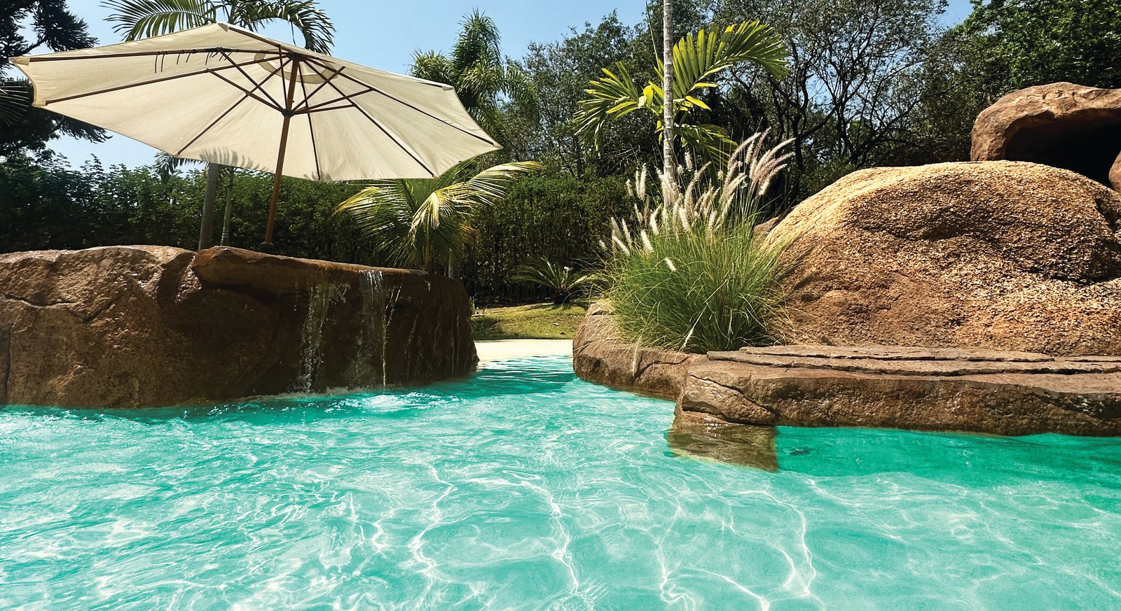 Piscina com pedras tematizadas, guarda-sol e água esverdeada sobre o revestimento de areia