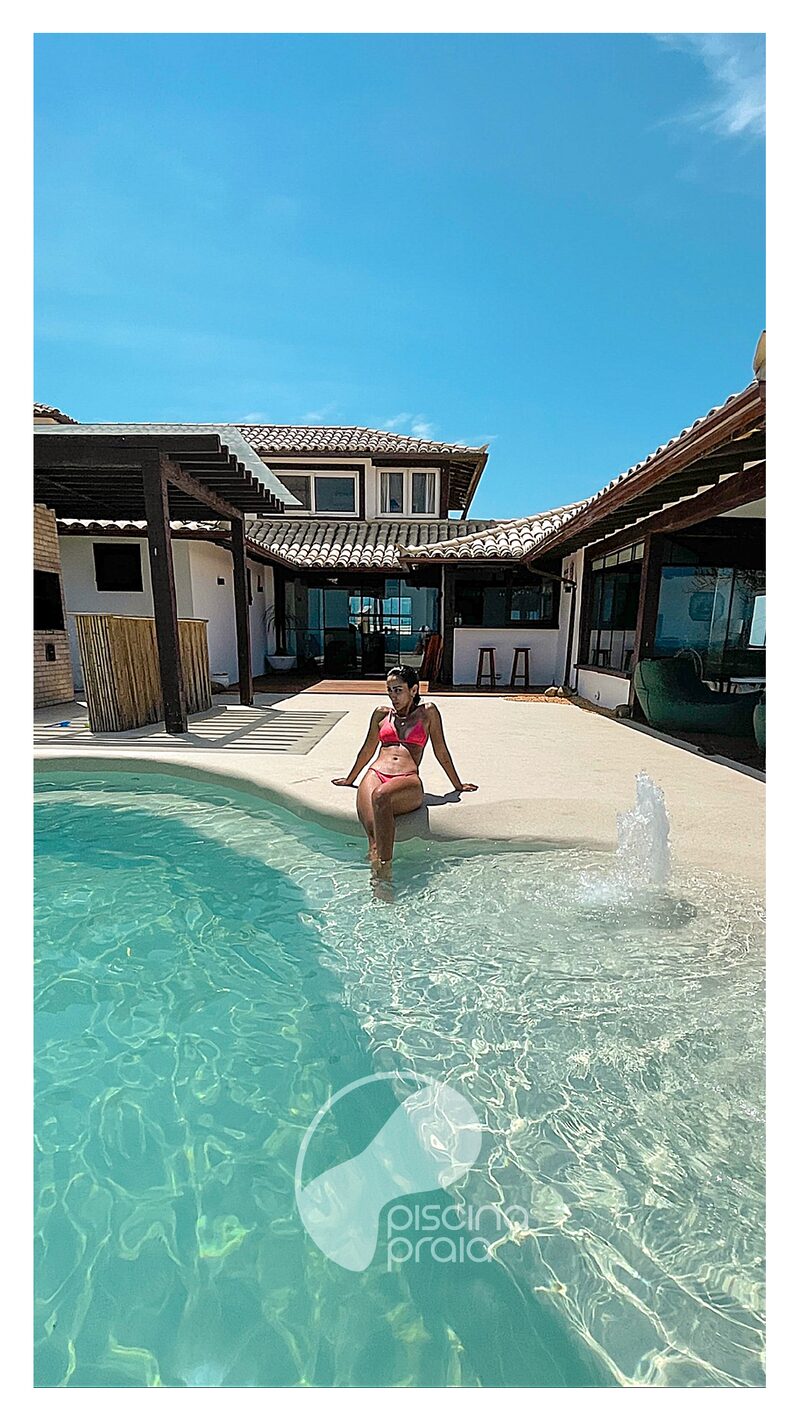 Piscina com prainha de areia em frente à casa, com pessoa sentada na borda