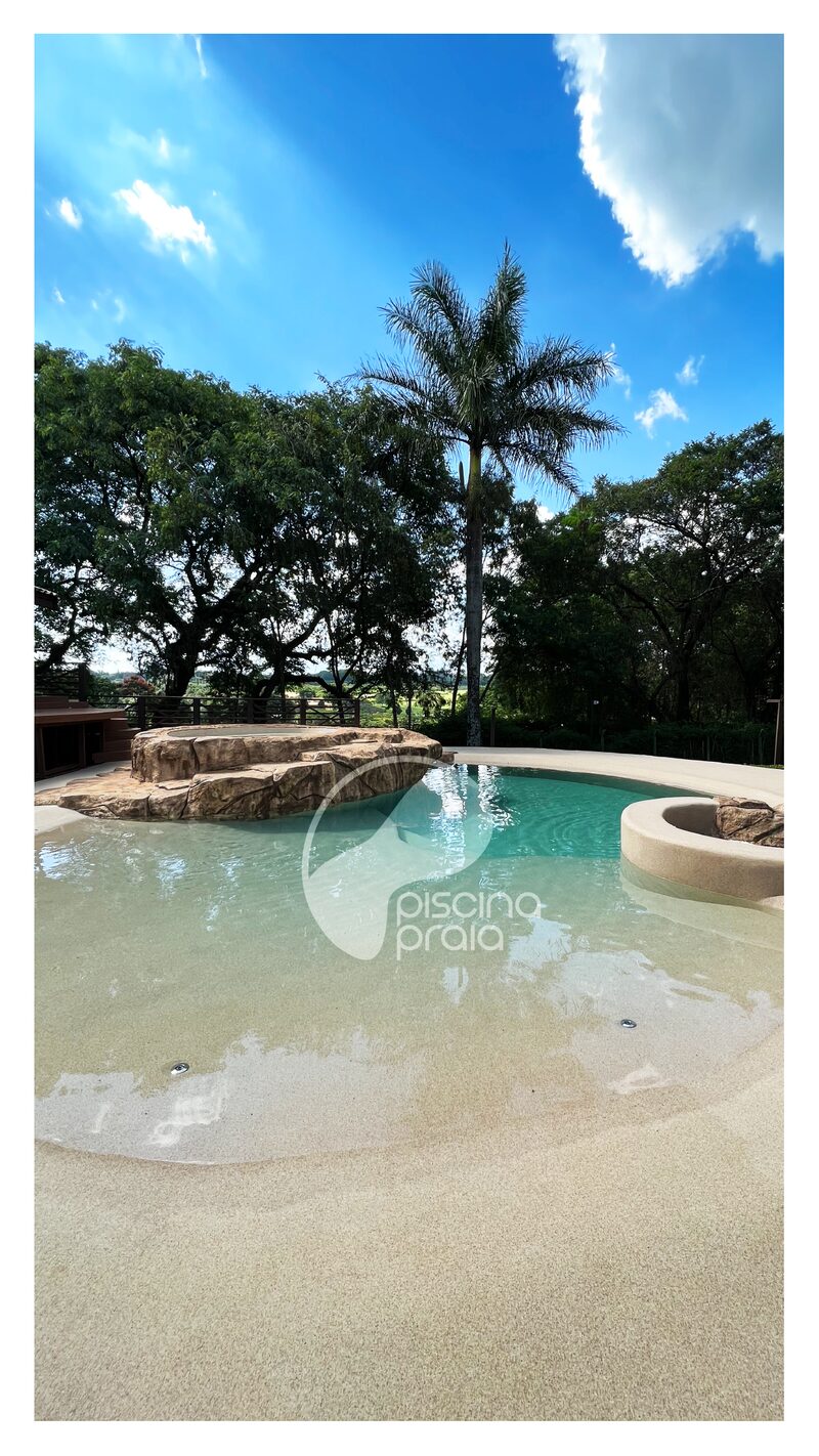 Piscina com prainha larga de areia, rochas e árvores ao fundo